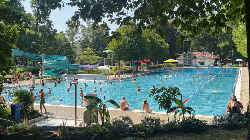 Das Bild zeigt das Schwimmbecken im Freibad Bittenfeld mit vielen Badegästen im und ums Becken. Links sieht man die Rutsche und im Hintergrund die grüne Liegewiese.