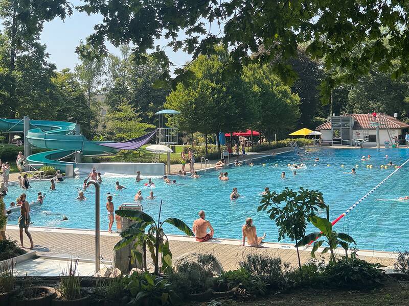 Das Bild zeigt das Schwimmbecken im Freibad Bittenfeld mit vielen Badegästen im und ums Becken. Links sieht man die Rutsche und im Hintergrund die grüne Liegewiese.