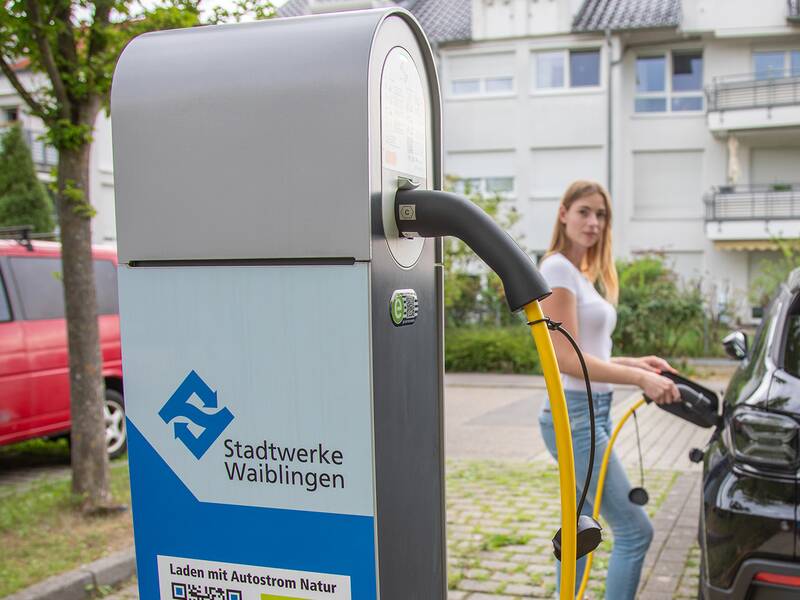 Das Stadtwerke Waiblingen-Logo auf einer Ladesäule steht im Fokus. Im Hintergrund lädt eine Junge Frau Ihr Auto.s
