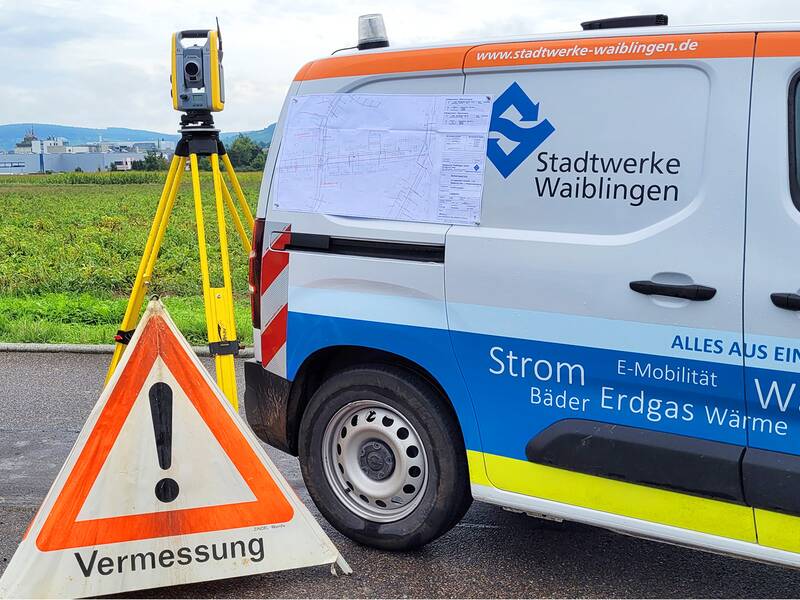 Ein Baustellenfahrzeug der Stadtwerke Waiblingen steht vor einem Feld. Neben dem Auto ist steht ein Messgerät und ein Warnaufsteller mit der Aufschrift "Vermessung".