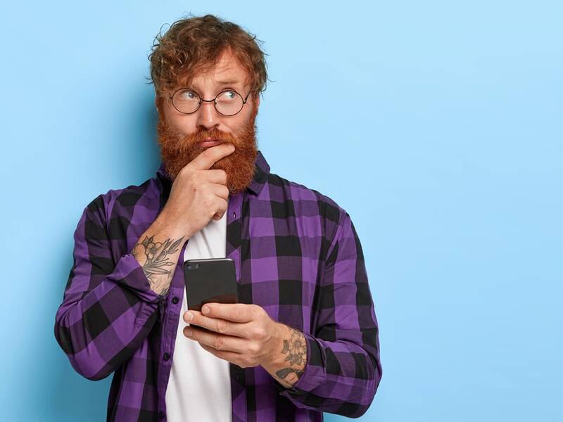 Ein Junger Mann min roten Harren und Vollbart in einer Überlegungspose mit einem Handy in der Hand