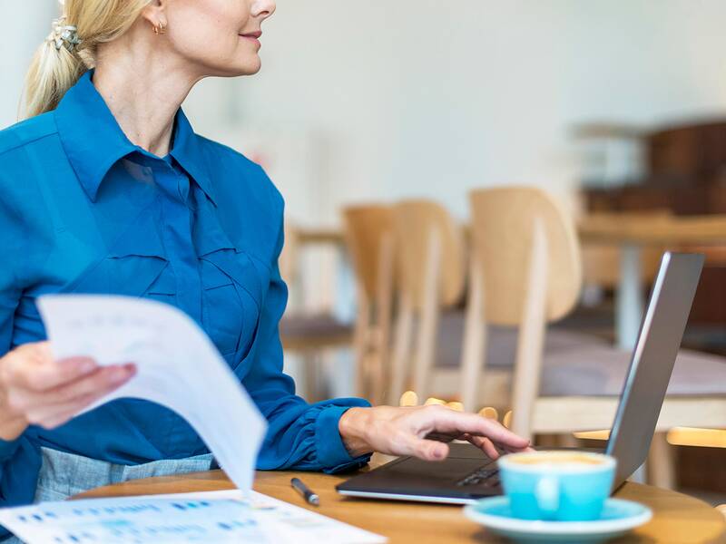 Eine Frau im blauen Outfit, die mit der einen Hand am Laptop arbeitet und in der anderen Hand ein Papier hebt. 