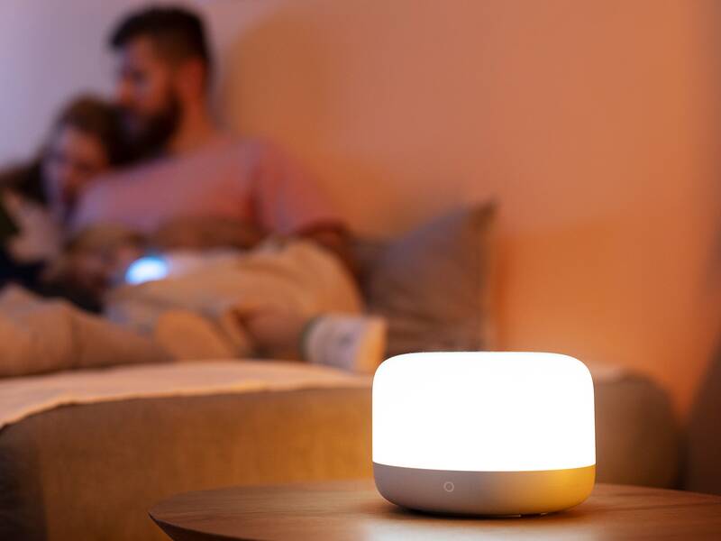 Das Bild zeigt im Vordergrund eine hell erleuchtete Tischlampe und im Hintergrund unscharf ein Paar, das sich gemütlich auf dem Sofa zusammengekuschelt hat
