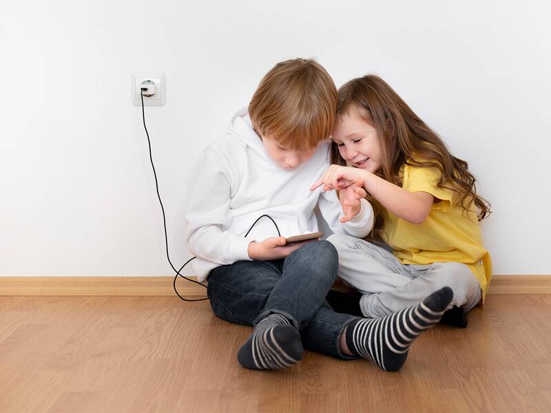 Zwei Kinder sitzen an einer Wand und spielen mit einem Handy das mit einem Kabel an einer Steckdose angesteckt ist