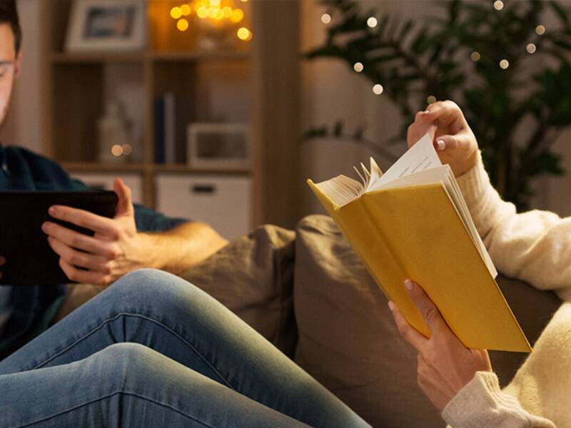 Mann und Frau auf dem Sofa zuhause. Die Frau liest ein Buch, der Mann ist am Tablet.