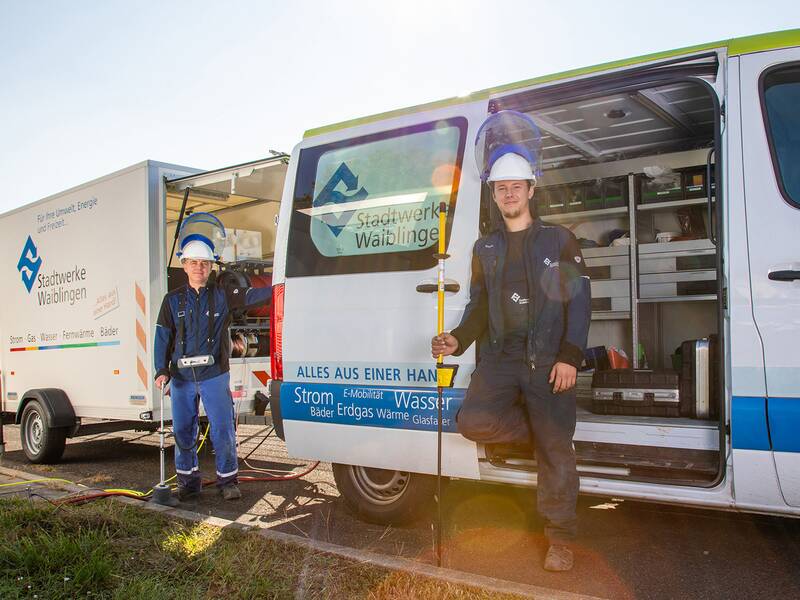  Das Bild zeigt zwei Arbeiter in blauer Arbeitskleidung und weißen Helmen vor einem geöffneten Lieferwagen der Stadtwerke Waiblingen. Sie halten Werkzeuge in den Händen und blicken in die Kamera. Im Hintergrund ist ein sonniger Tag mit etwas Grünfläche zu sehen. Der Lieferwagen trägt das Logo und die Dienstleistungen des Unternehmens.