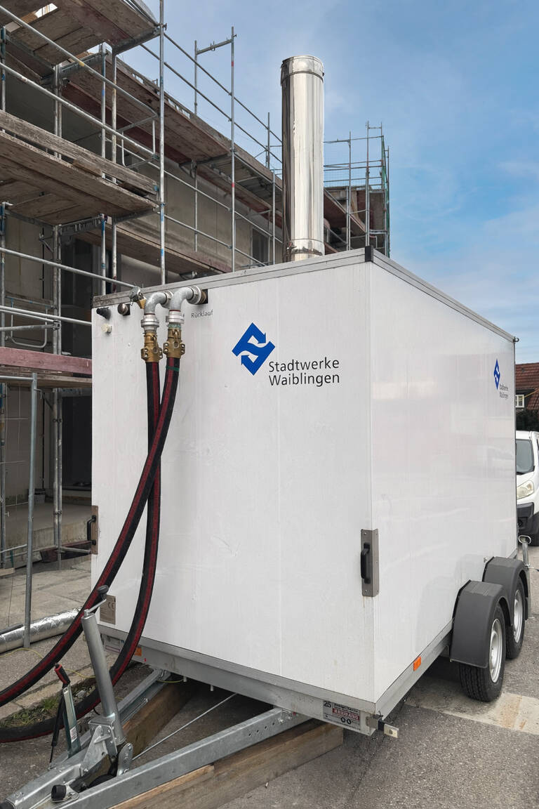 Das Bild zeigt die mobile Heizzentrale, ein weißer Kastenförmiger Anhänger mit Anschlüssen und Stadtwerke Waiblingen Logo. Die Heizzentrale ist angeschlossen und steht vor einer Baustelle mit Gerüst.